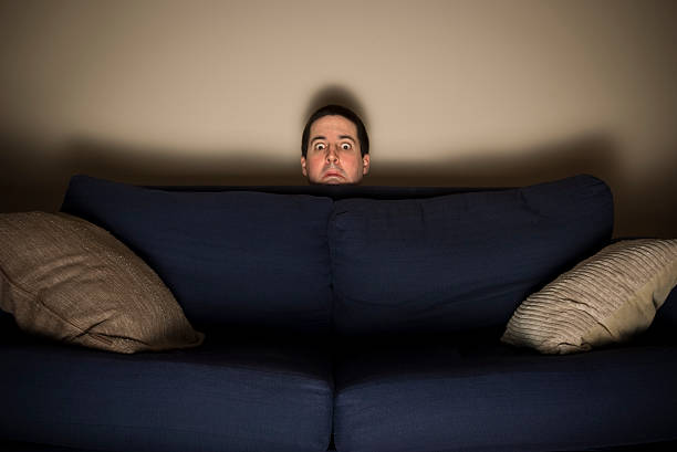 frightened-man-peeks-over-a-couch-while-watching-tv-picture-id176773021